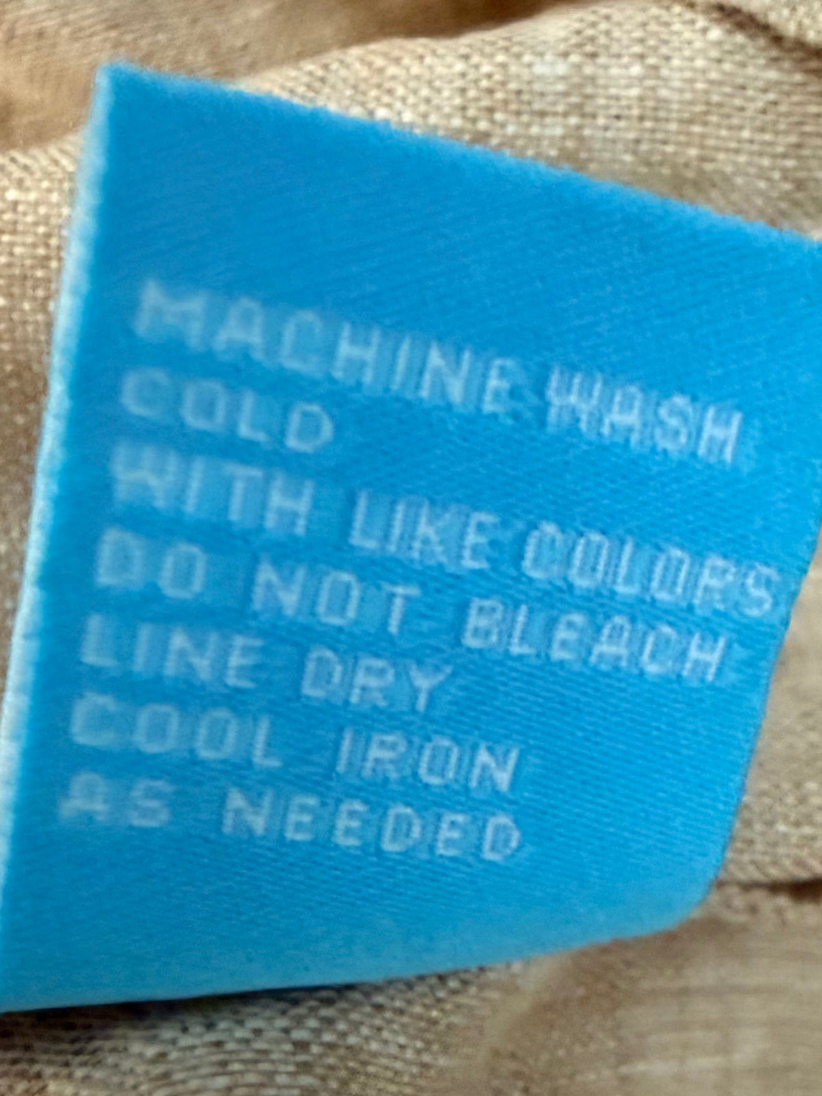 A close-up of a beige woven fabric garment with a light blue sewn-in care label reading 'MACHINE WASH COLD WITH LIKE COLORS DO NOT BLEACH LINE DRY COOL IRON AS NEEDED.'.